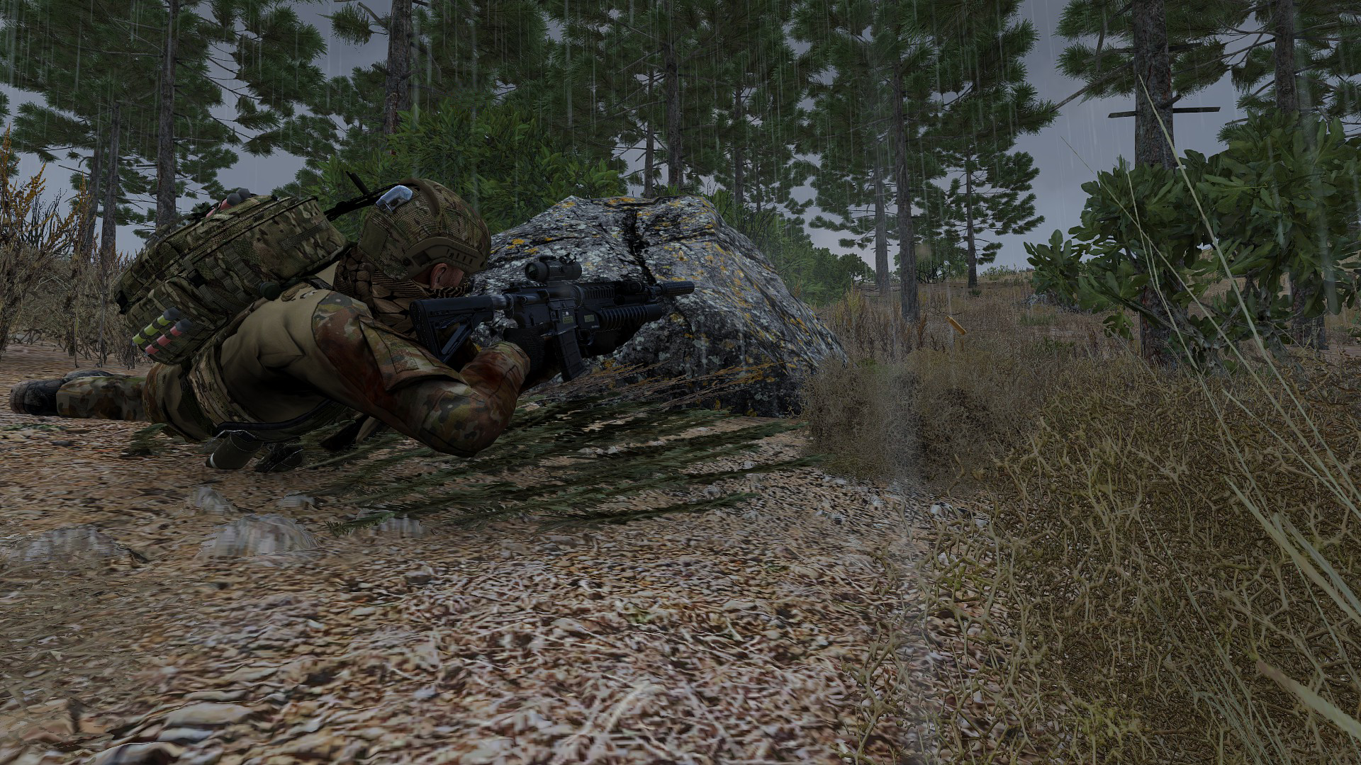 A soldier in camouflage gear lying prone on the ground aiming a rifle in a forested area during rainy weather.