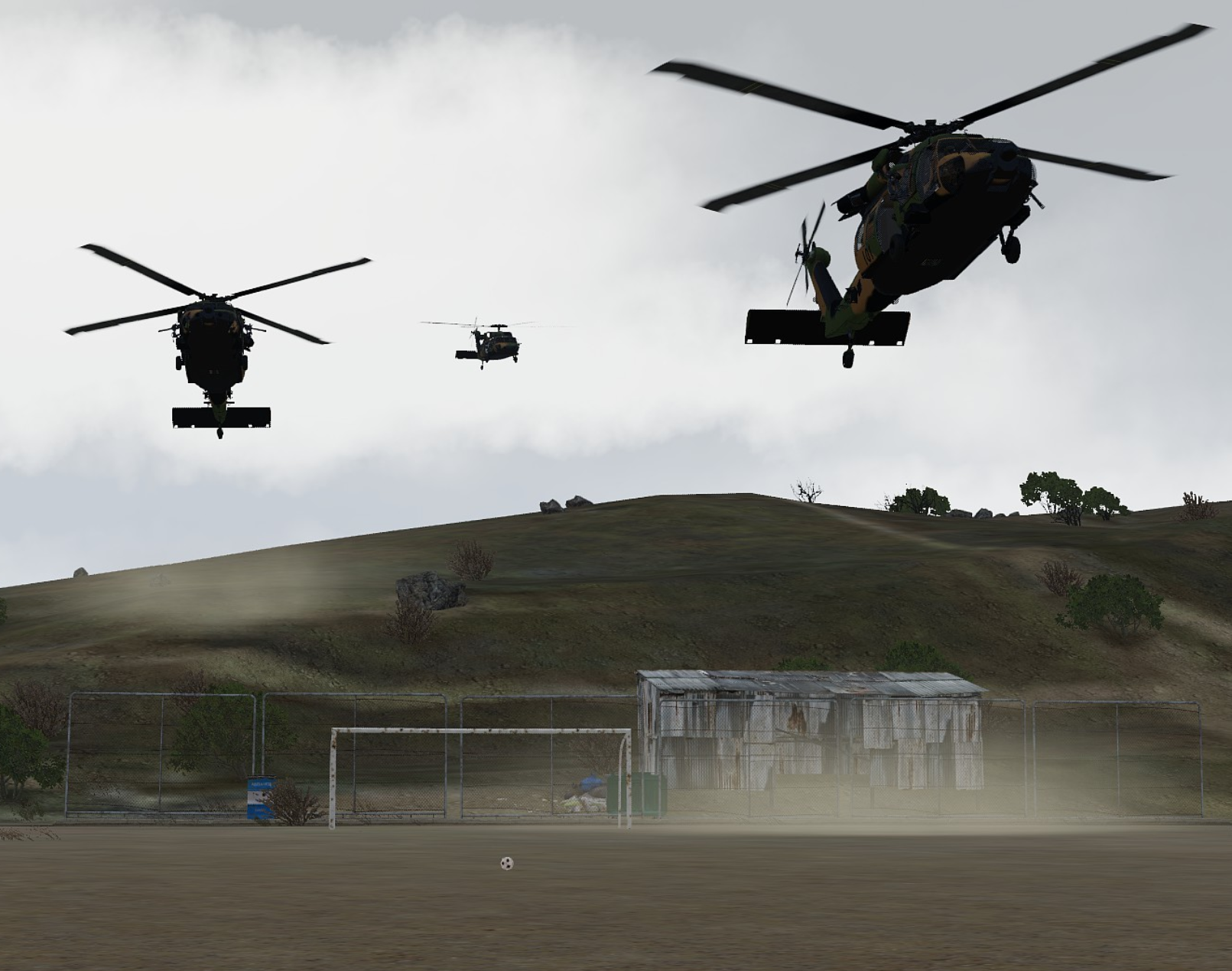 Three military helicopters flying over a soccer field with a goal, a rusty shed, and a hill with trees in the background.