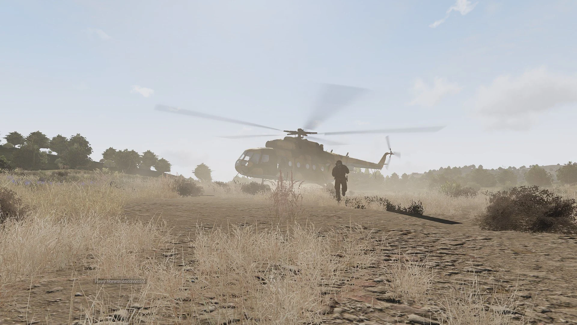 A military helicopter flying low over a dirt path with a soldier running towards it in a dry, grassy landscape.